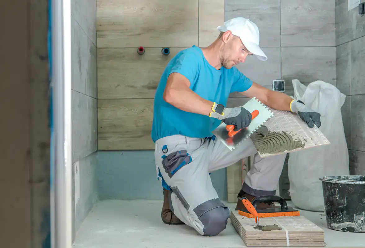 A man in work clothes applies adhesive to a tile in a Suffolk County bathroom under construction.