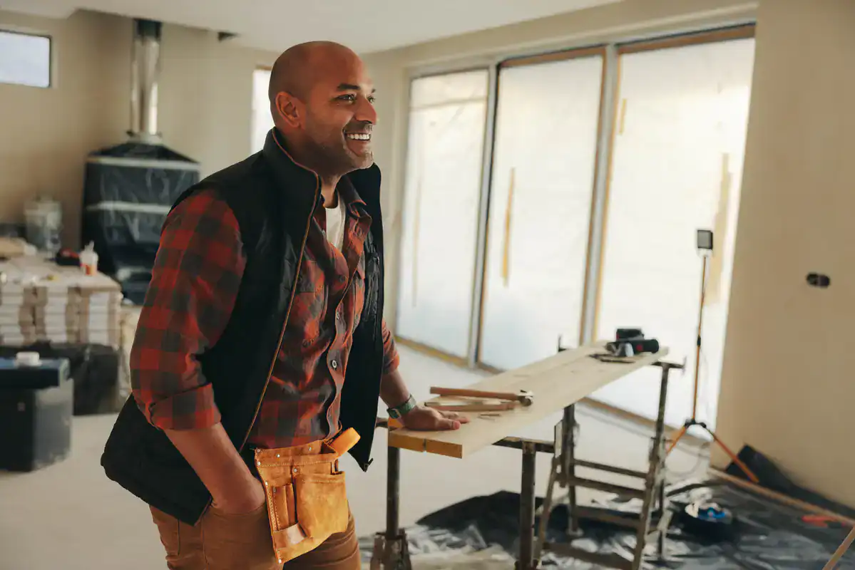 Smiling man in flannel and tool belt stands by wood and tools, ready for Suffolk County countertop install.