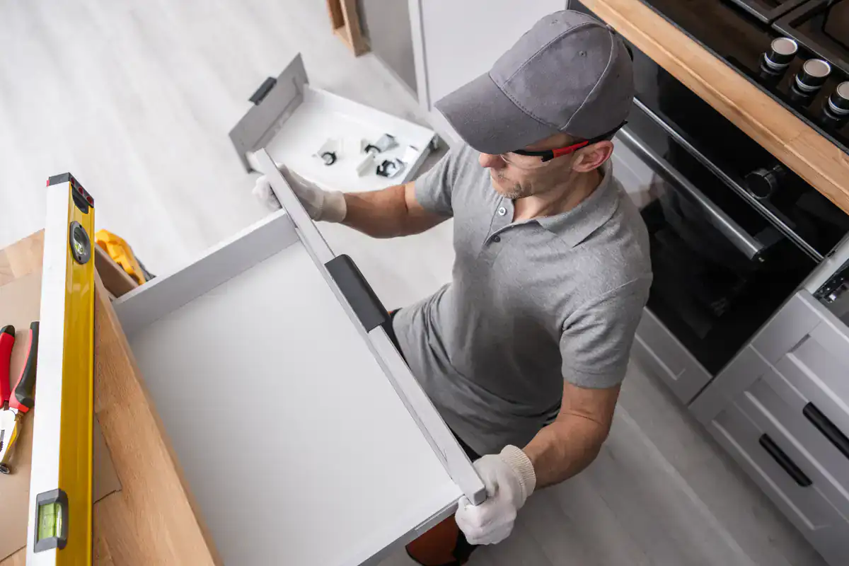A man in gloves installs a kitchen drawer front in a modern NY kitchen with cabinets and tools nearby.